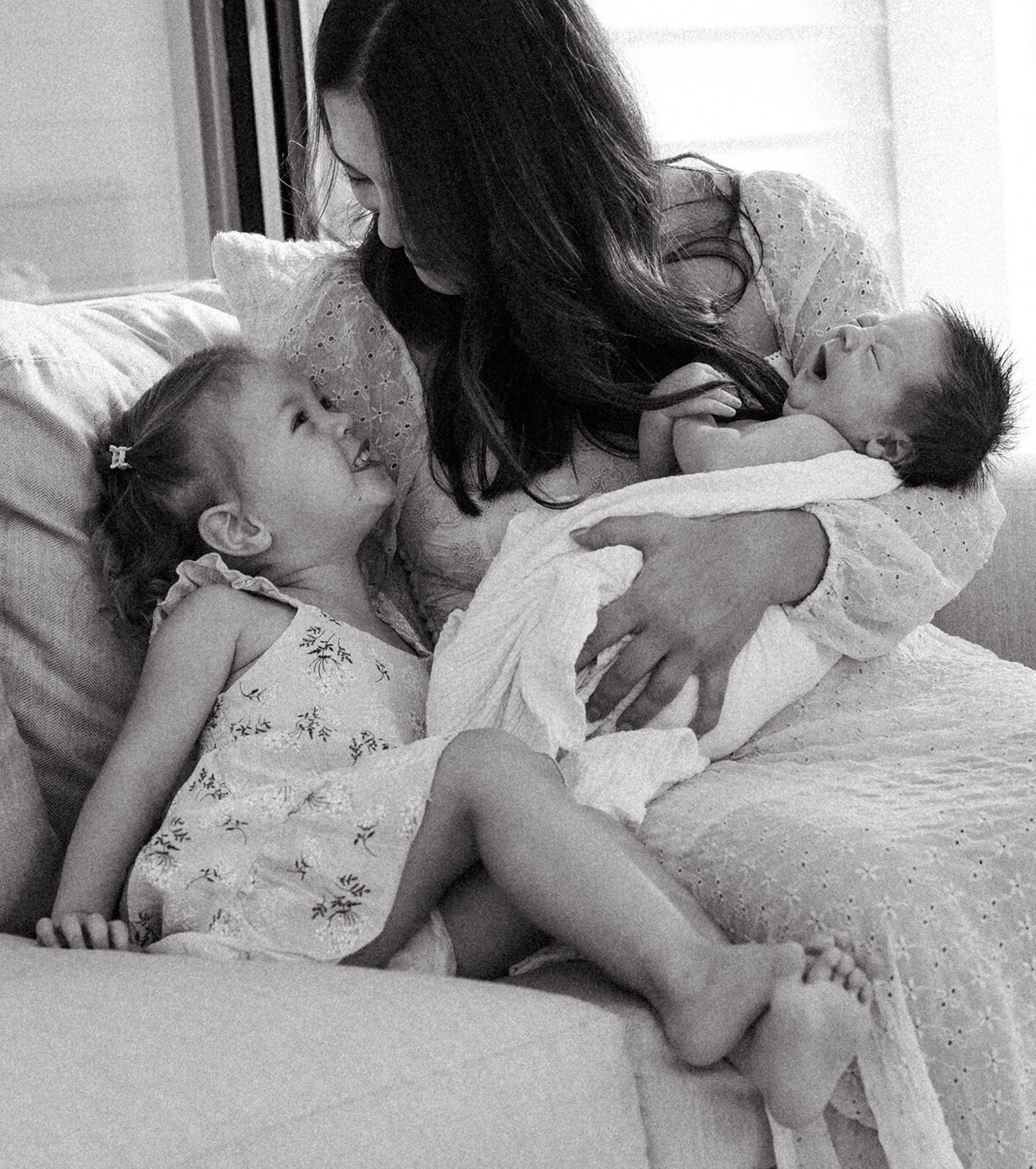 Toddler girl smiling up at her mother, who is holding a newborn, during an in-home family photo session