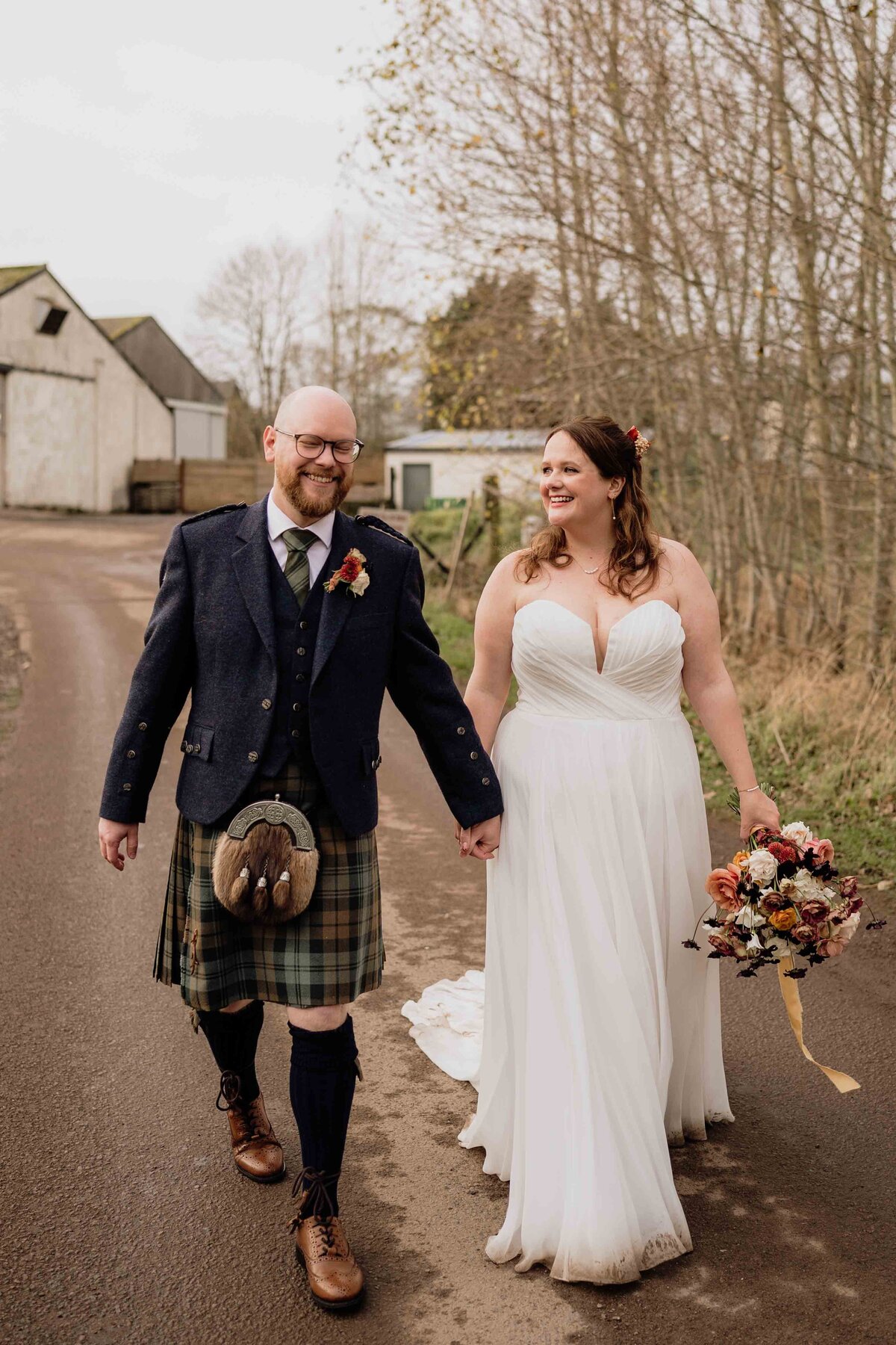 A couple is walking hand in hand on a rustic path at Charleton Fruit Farm. Captured by an Aberdeen wedding photographer, the groom wears a kilt and jacket, while the bride dazzles in a white dress with a bouquet. They smile at each other, surrounded by bare trees and old buildings.