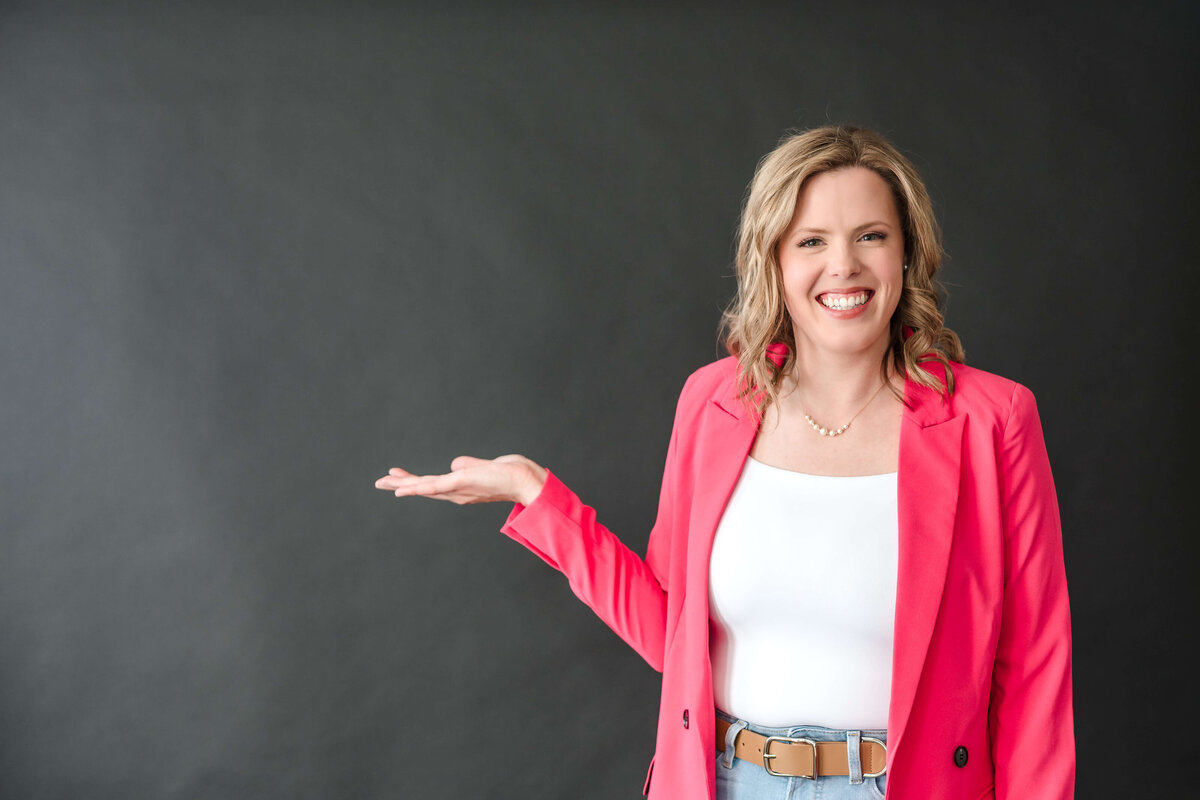 Female entrepreneur smiling and gesturing with open palm while wearing pink blazer.