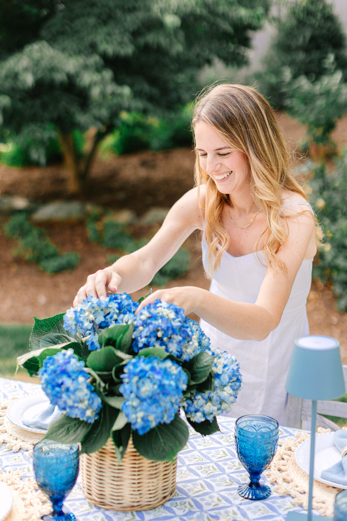 A smiling woman in a white sundress arranges a bouquet of vibrant blue hydrangeas in a wicker basket on an outdoor dining table. The table is set with blue glassware, woven placemats, and a patterned tablecloth, creating a charming garden setting