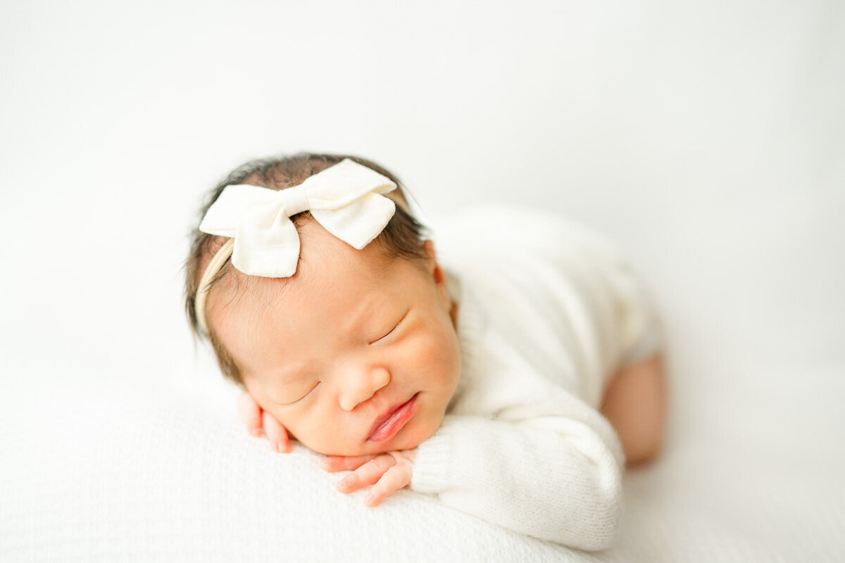 a newborn girl is posed on a white bean bag. She lays on her stomach and has her hands tucked under her chin while softly smiling. 