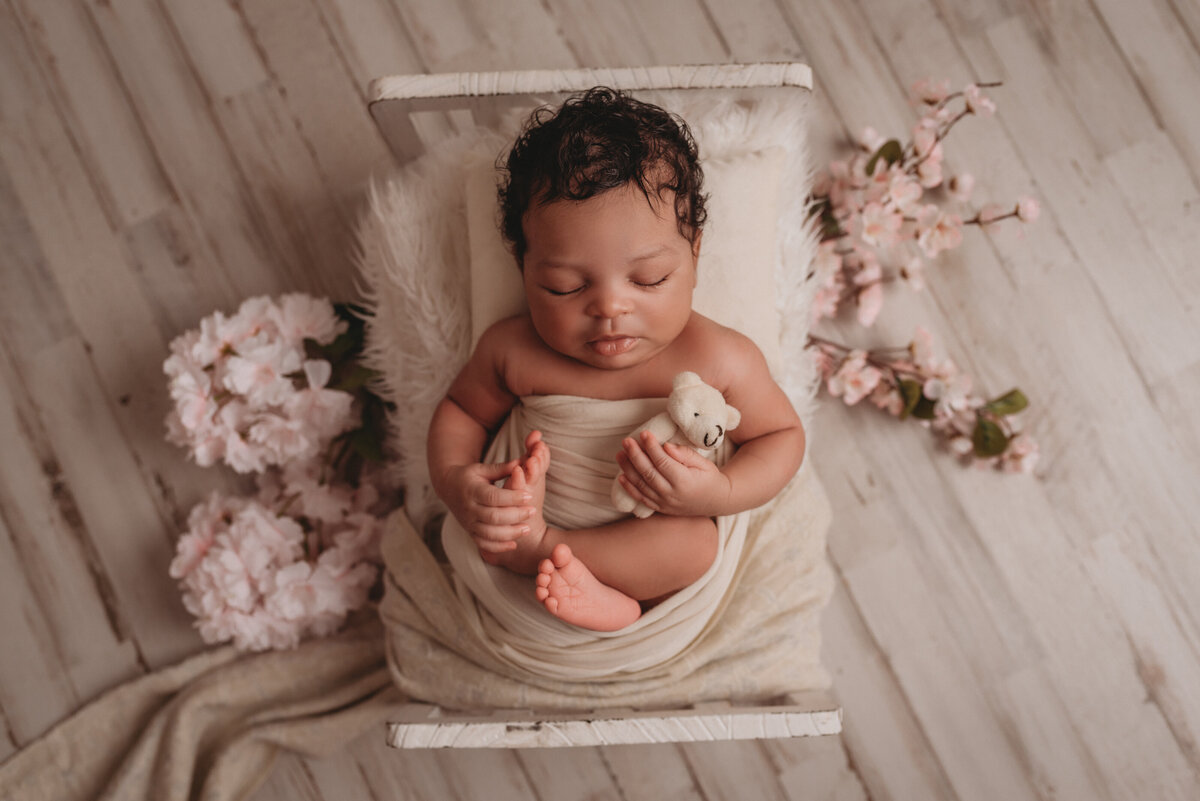 Newborn girl asleep and wrapped in white fabric in miniature white bed on white wood floor backdrop