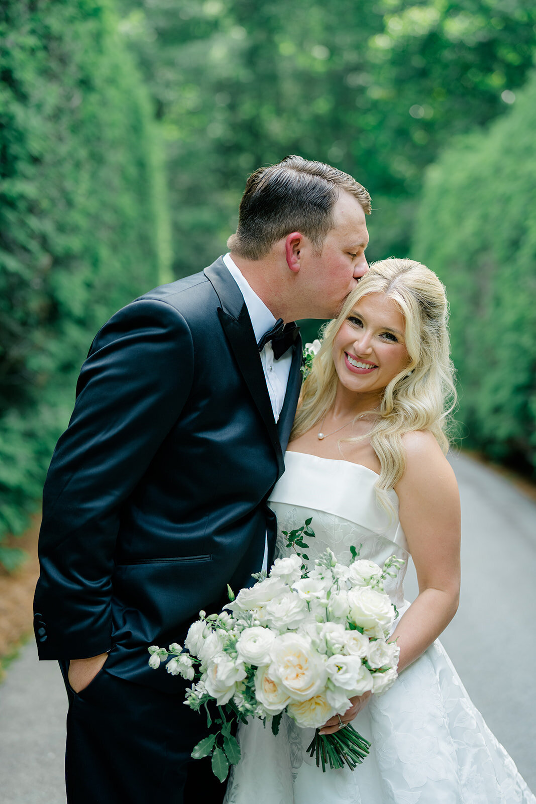 Close-up portrait of groom kissing bride’s forehead while holding a white rose bouquet outdoors.