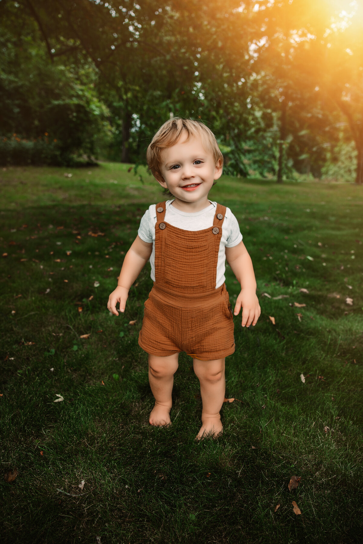 toddler in overalls smiles at the camera at sunset in front yard