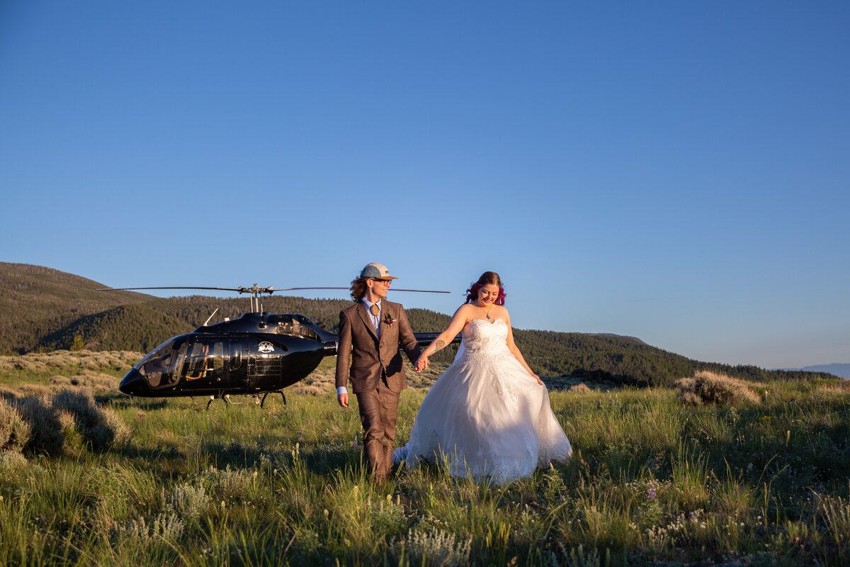 A bride and groom walk hand in hand through a field with a helicopter parked behind them.