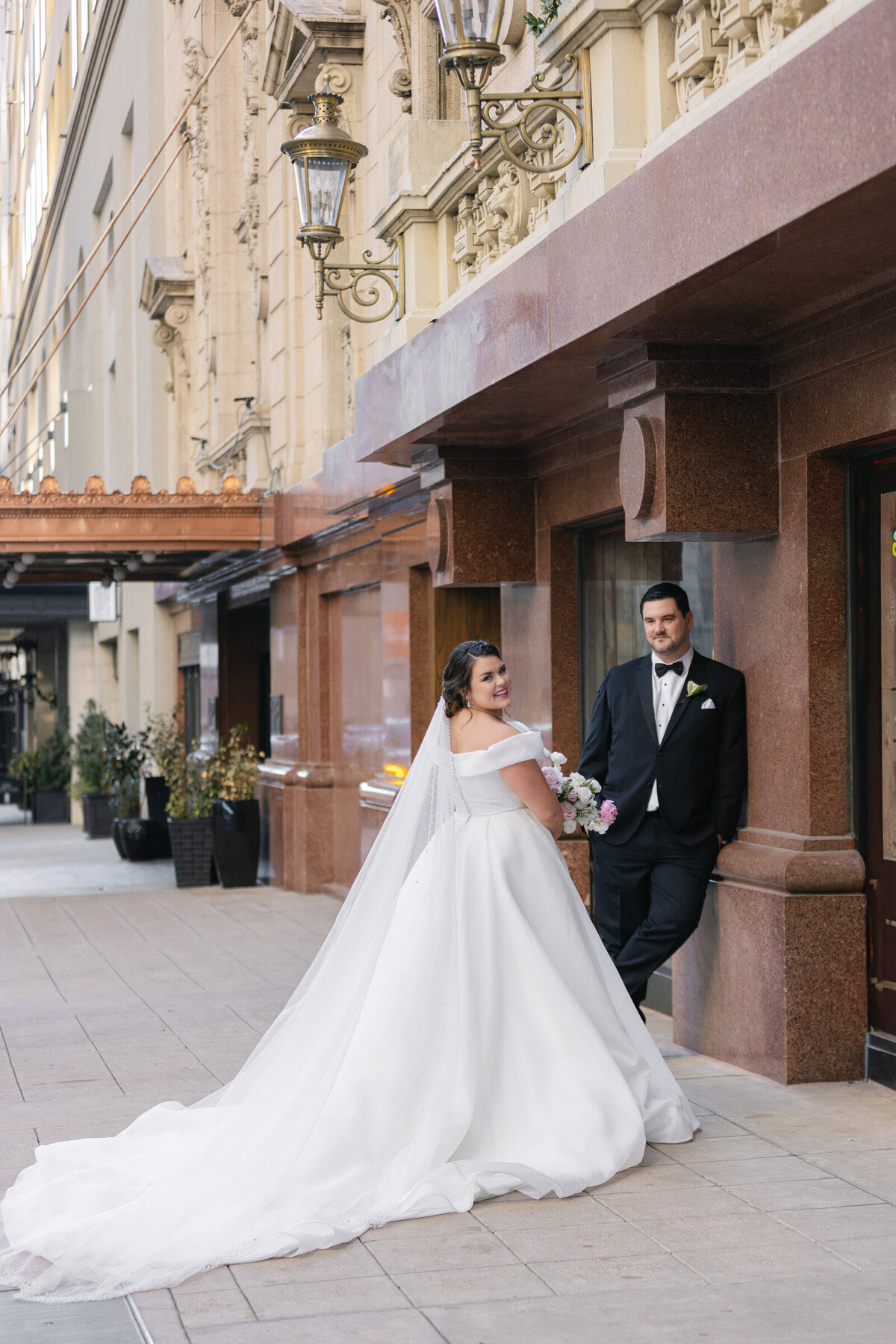 bride and groom outside The Adolphus in Dallas, with the groom leaning on a wall looking at the bride while she faces the camera, capturing a stylish and romantic wedding portrait.