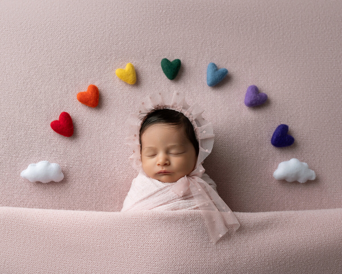 Rainbow baby girl in soft pink wrap and bonnet surrounded by colorful hearts and clouds