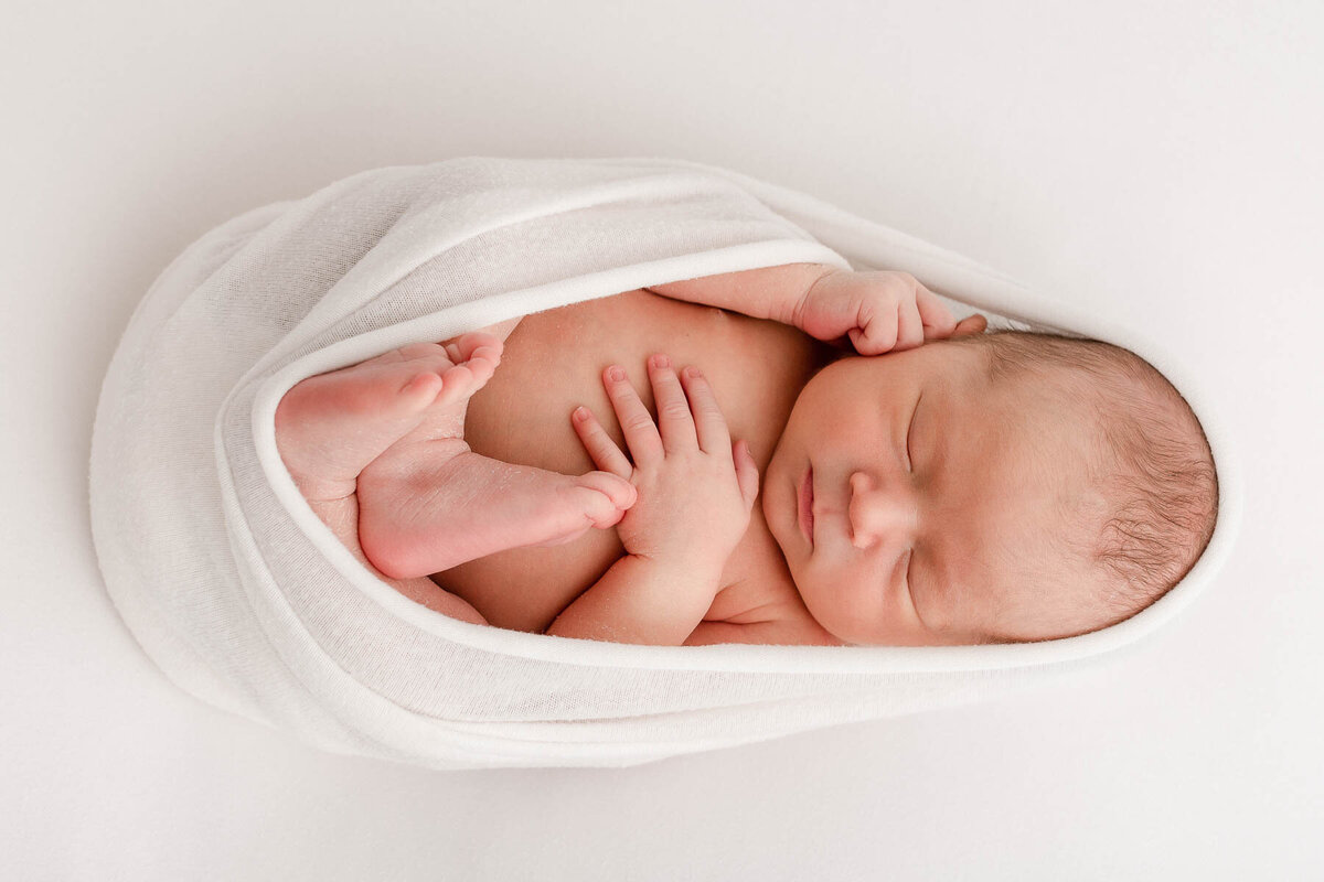 Sleeping baby swaddled in white laying on white backdrop with fingers and toes showing. Newborn Photo Session.