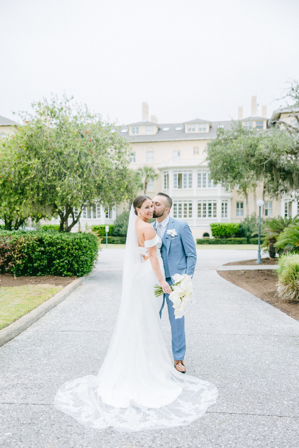Groom kisses his bride’s cheek while strolling the tree-lined path outside the historic Jekyll Island Club Resort—captured by luxury wedding photographer Amia Marcell.