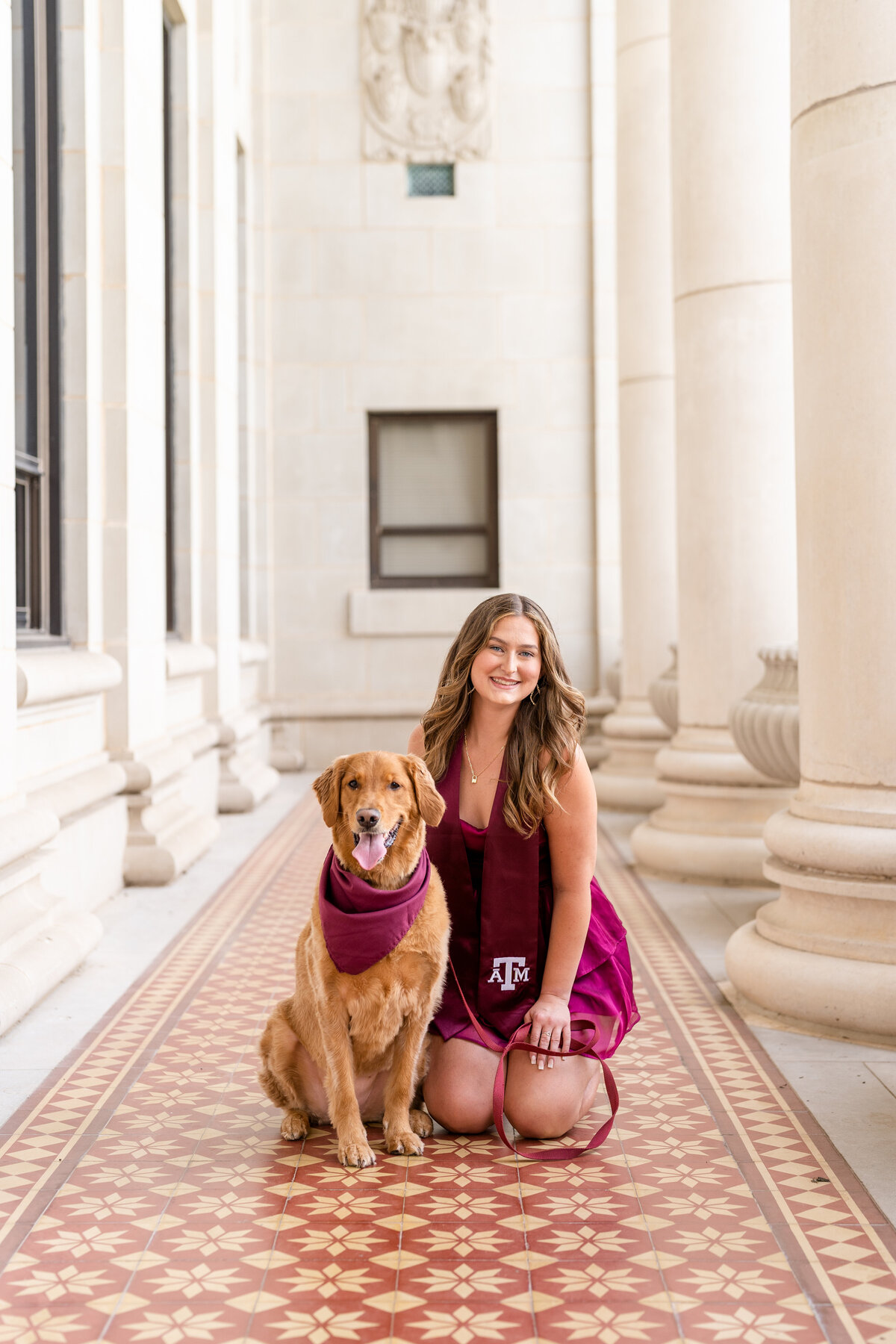 Texas A&M senior girl kneeling next to dog while wearing maroon dress and Aggie stole in the columns of the Administration Building