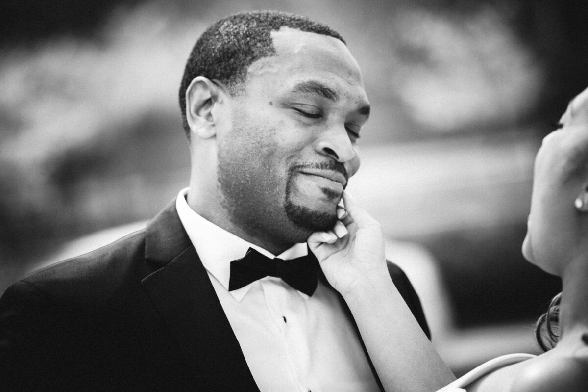 A groom smiling as a woman reaches up towards his face.