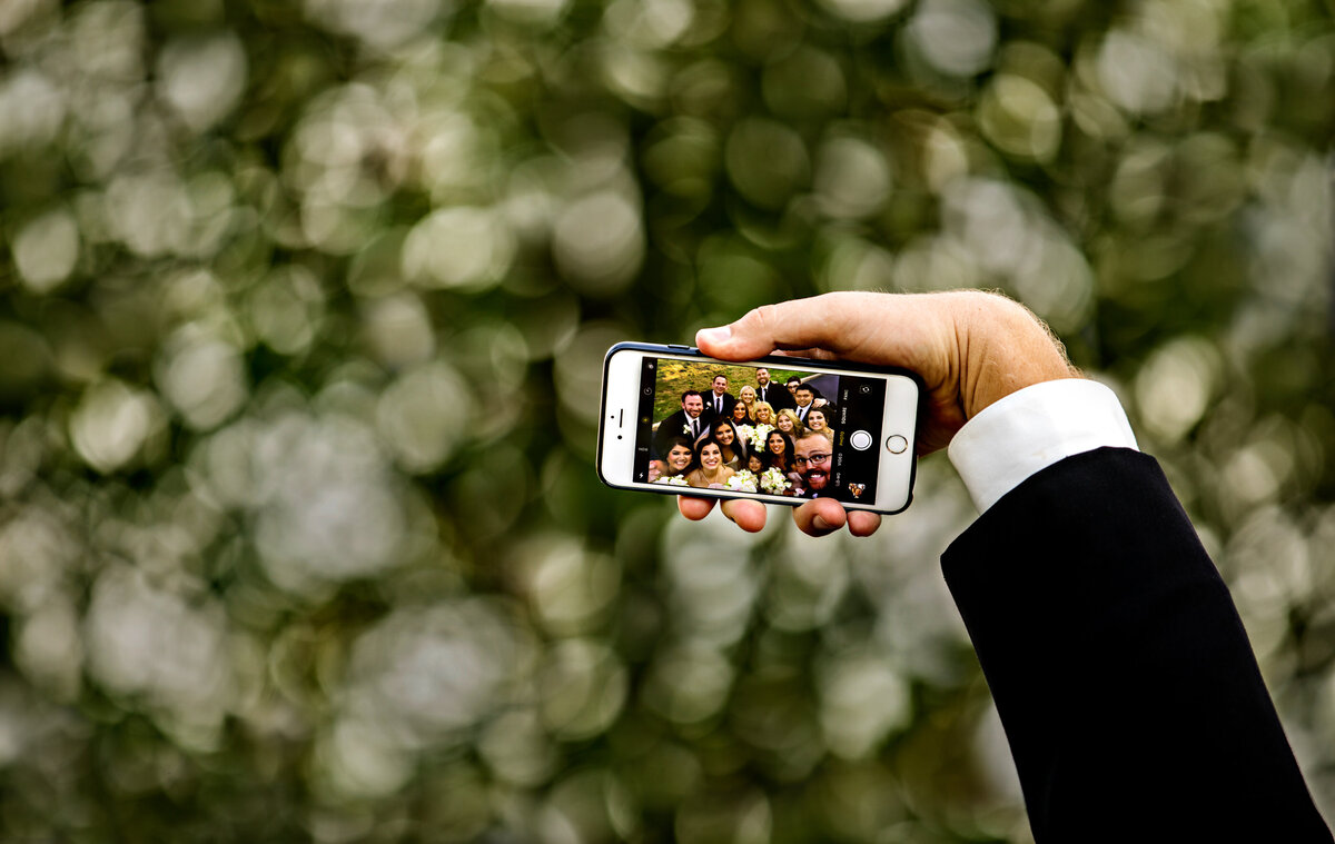A tall groomsman takes a selfie with the bridal party. His hand and phone are visible with the bridal party on the screen.