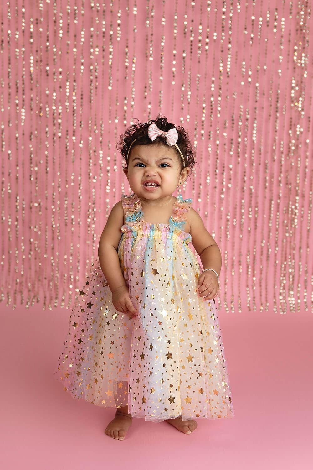 A young toddler girl in a rainbow princess dress makes a grumpy face while standing in a studio