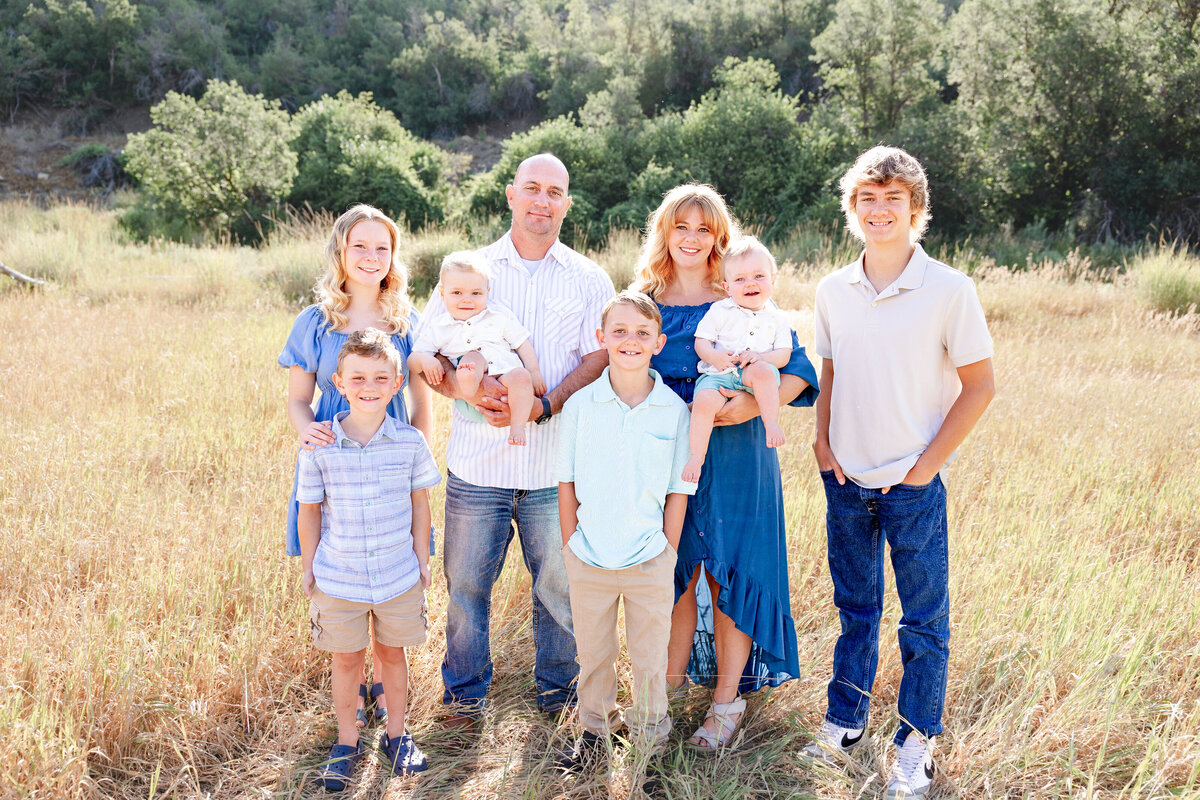 A large family posing in a sunlit field, dressed in coordinated blue and neutral tones, with a beautiful mountain backdrop.