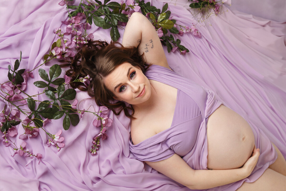 Maternity session of a beautiful young woman laying on purple fabrig with purple flowers taken by life in Pink Photography studio in Orfordville, Wisconsin