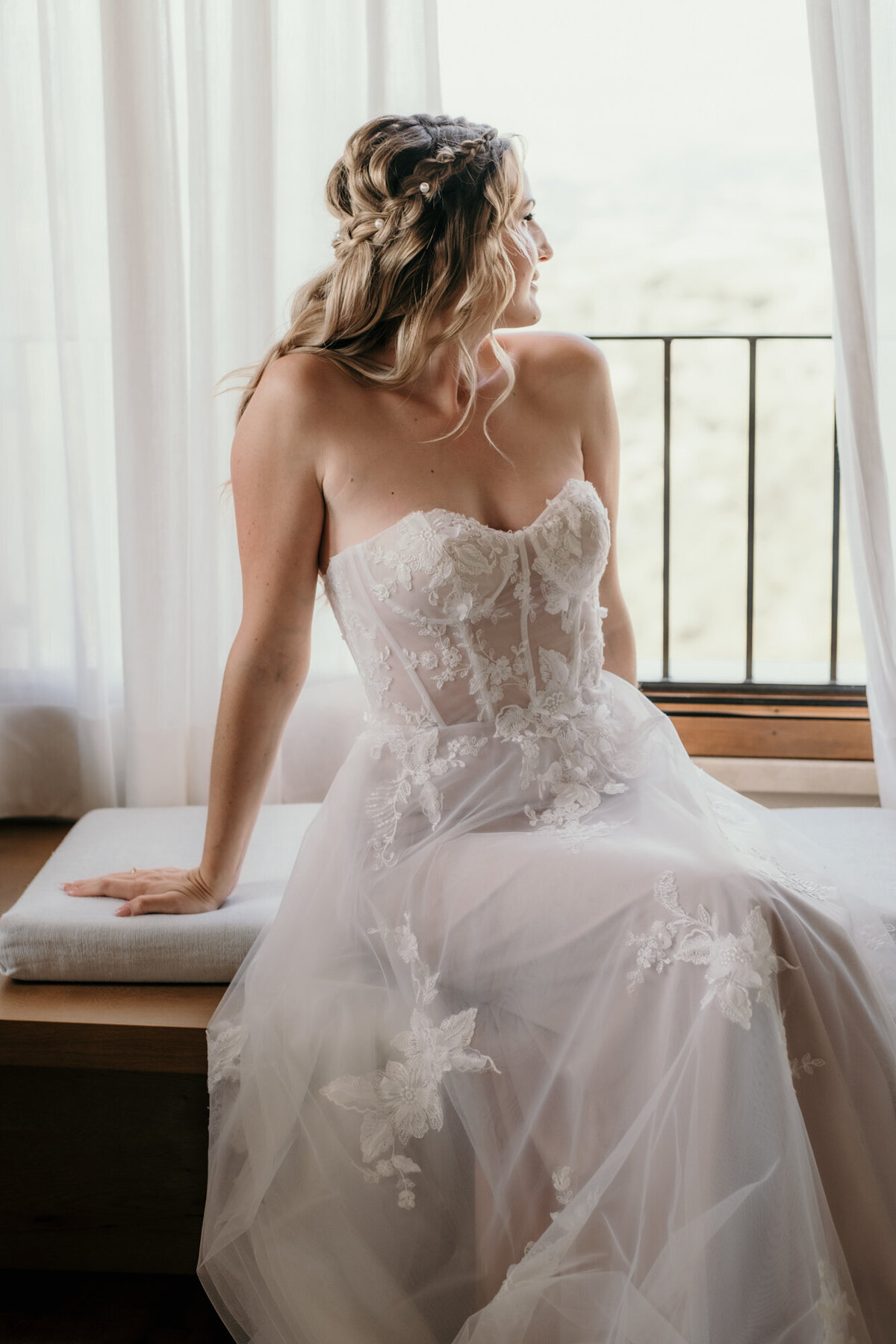 Die Braut sitzt fertig gestylt auf einem Fensterbrett und blickt verträumt aus dem Fenster.