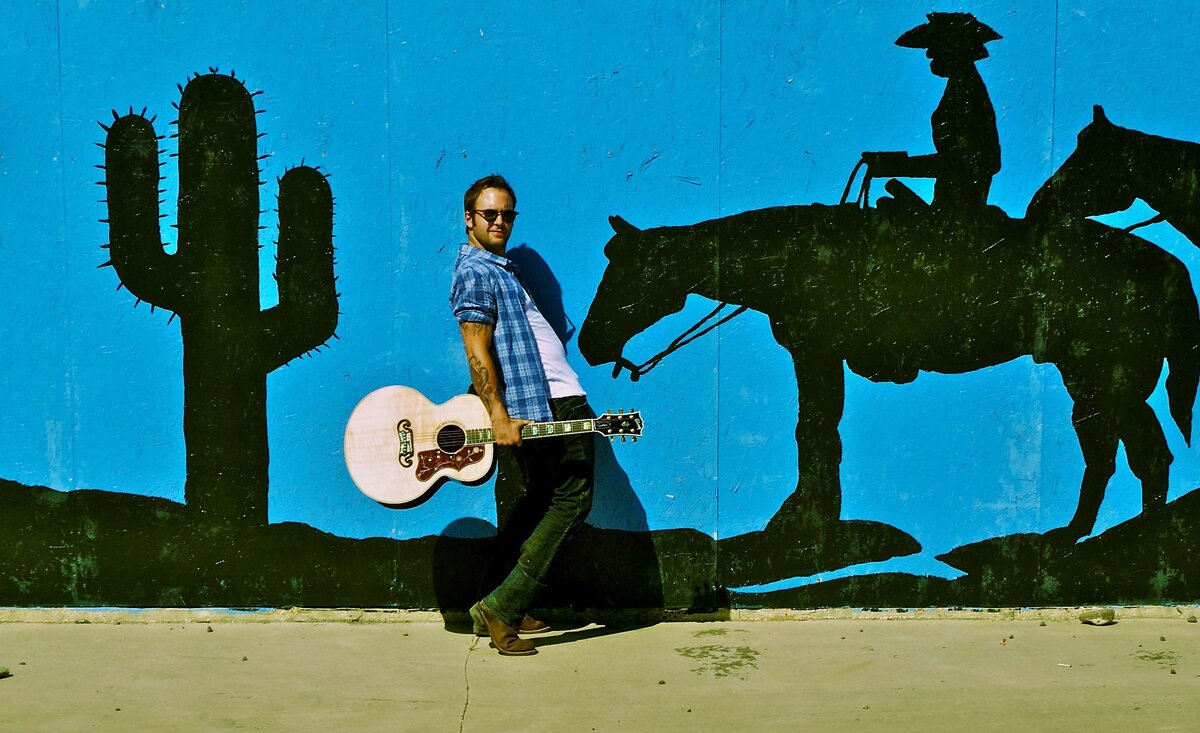Male musician photo Dallas Smith wearing blue plaid shirt holding tan guitar leaning against horse with cactus mural