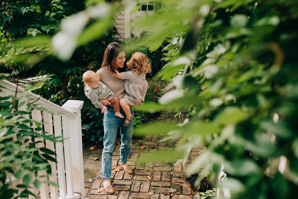 A mother wearing a striped shirt and jeans holds her two young daughters on her back patio