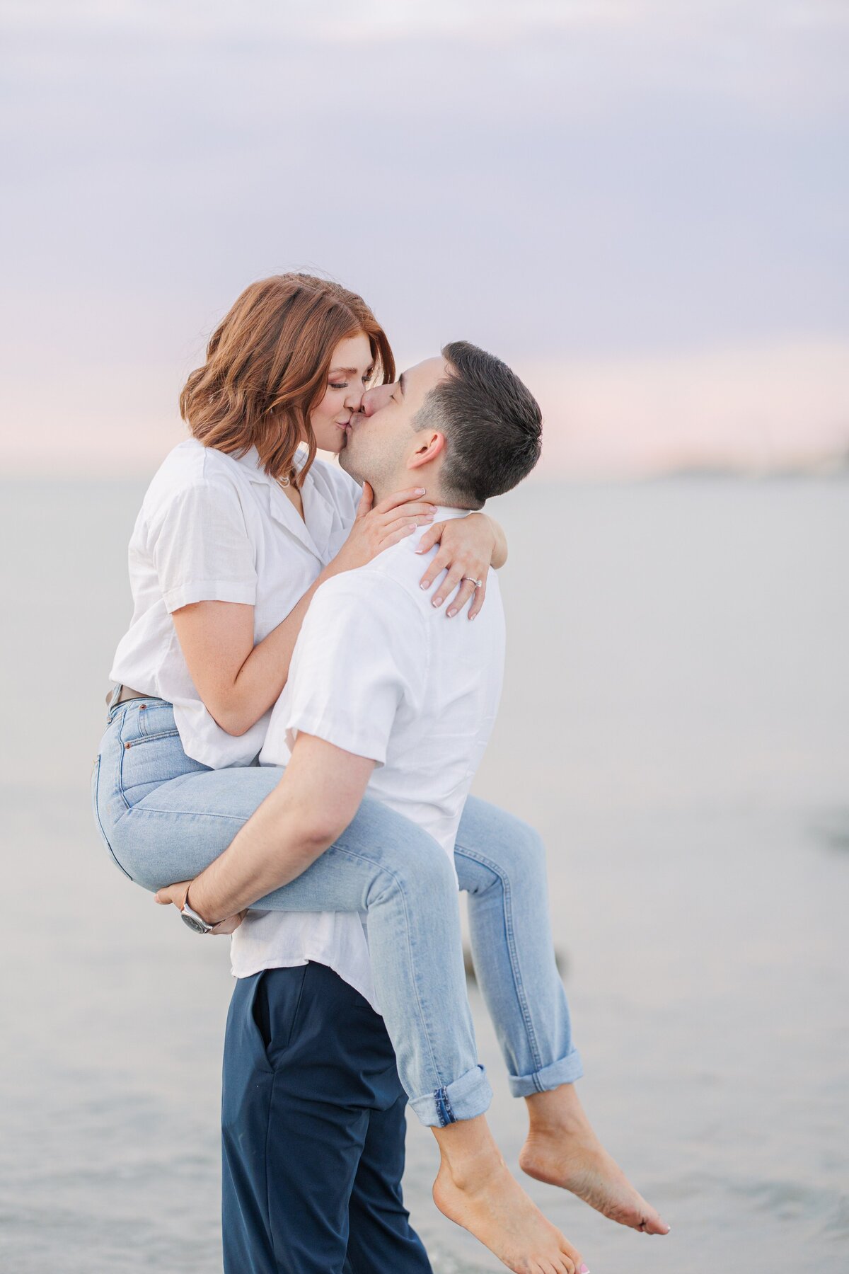 A man picking up and kissing a woman during their Wingaersheek Beach sunset engagement photography session.