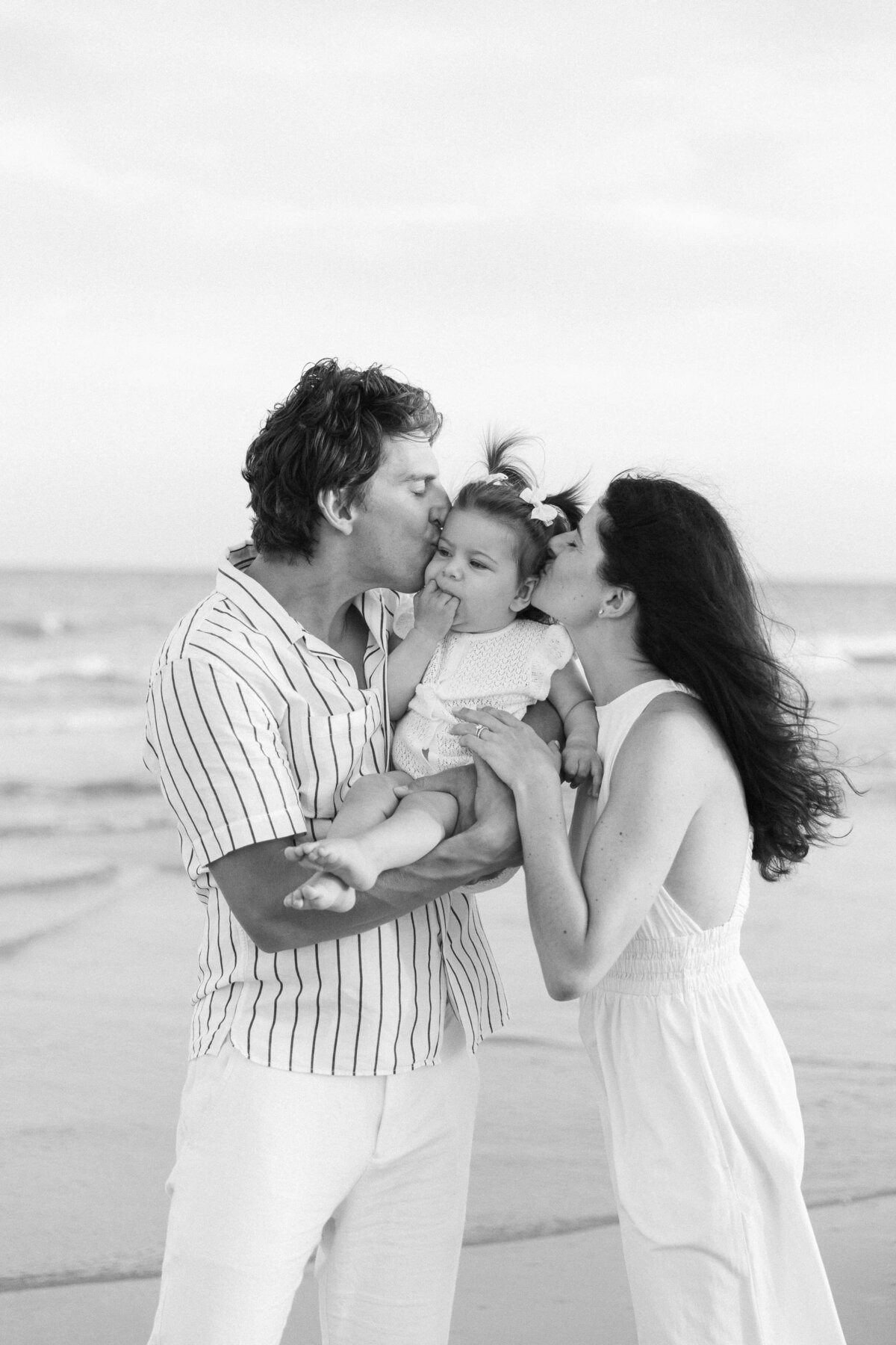 Mom and Dad kiss baby girl on her cheeks candidly on the beach for Hilton Head family photographer, Lamp and Light Photography.