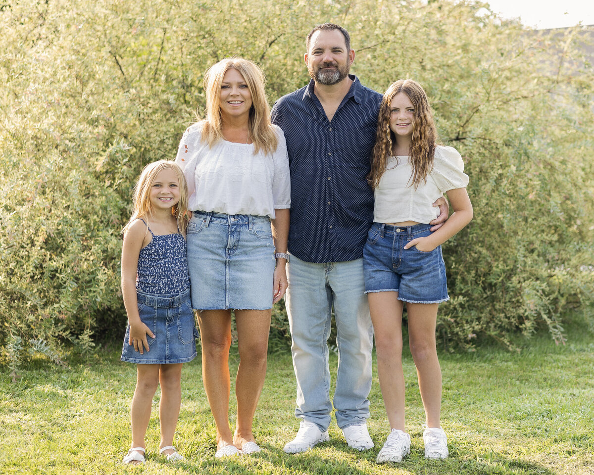 Mom, Dad, and daughters standing in the grass in front of a bush, looking at the camera smiling.  Older daughter is wearing blue jean shorts, a white short sleeve shirt, and has long brown hair.  Youngest daughter has a dark blue and white tank top style shirt with a blue jean skirt, and has medium length blonde hair.  Mom is wearing a jean skirt, white half sleeve shirt, and has long blonde hair.  Dad is wearing a blue button up short sleeve shirt, blue jeans, and has black gray short hair and beard.