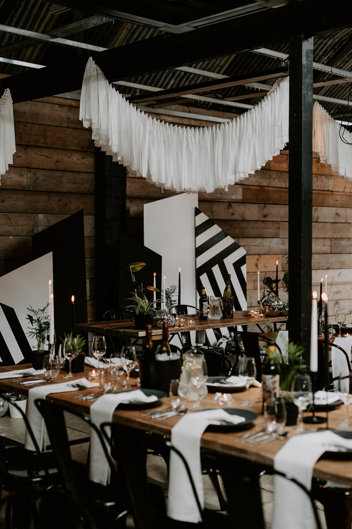 The Shack Revolution is set out for dinner with trestle tables, black and white boards are placed at a head table and black plates sit under white napkins. Plants are scattered around the room to give the space more green.