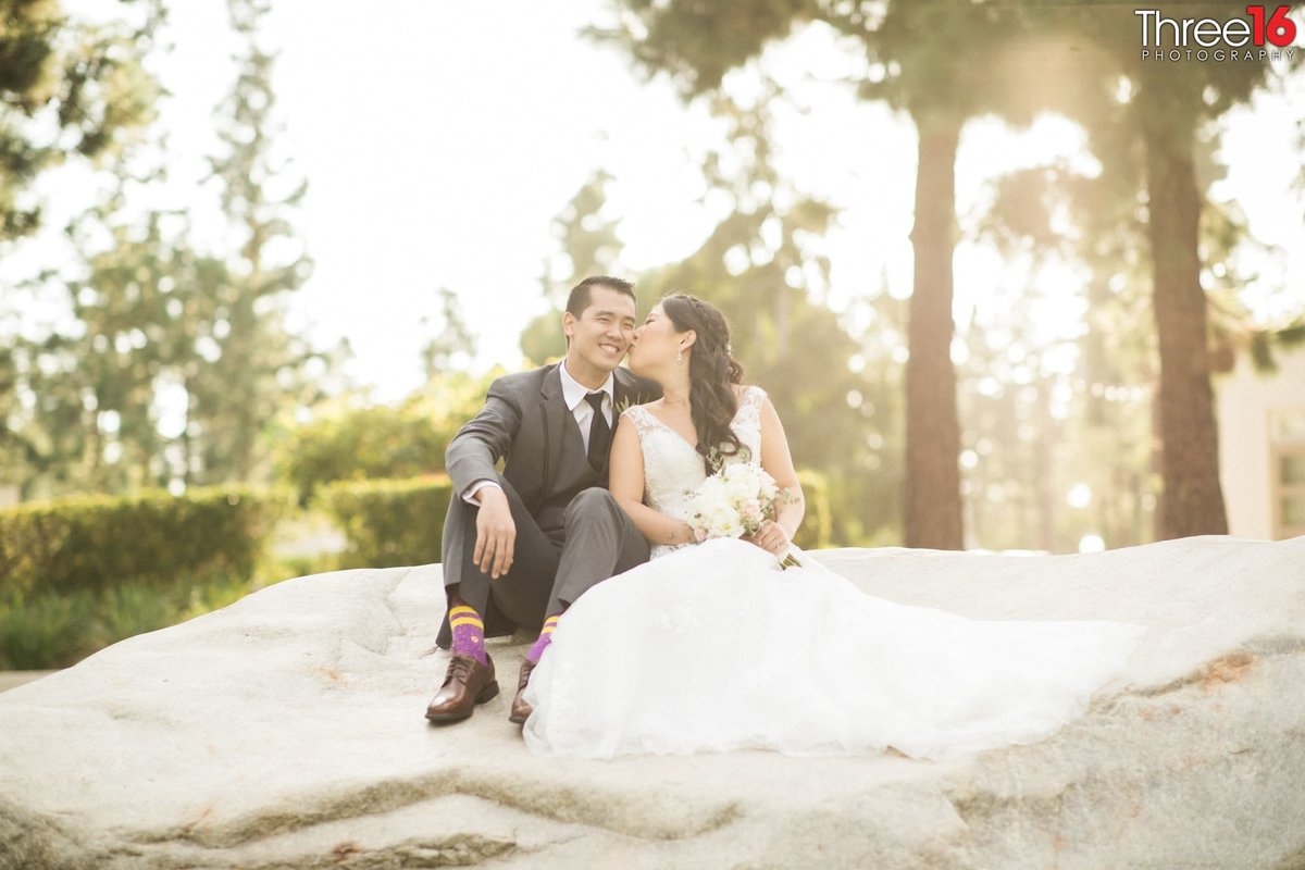 Bride kisses her Groom's cheek while sitting on a large rock