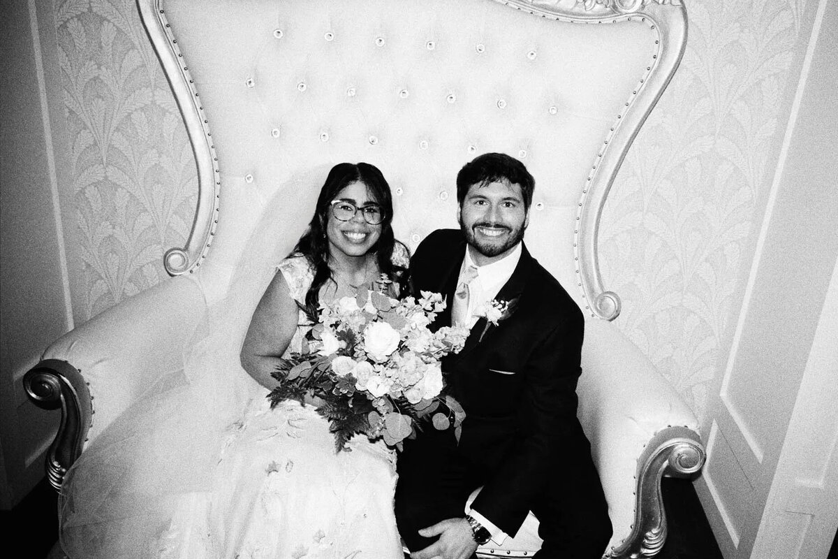 A bride and groom sit closely together on an ornate, high-backed chair. The bride holds a bouquet and smiles, while the groom, in a suit, sits beside her. Captured by a film photographer NJ, this black and white photo radiates timeless charm.