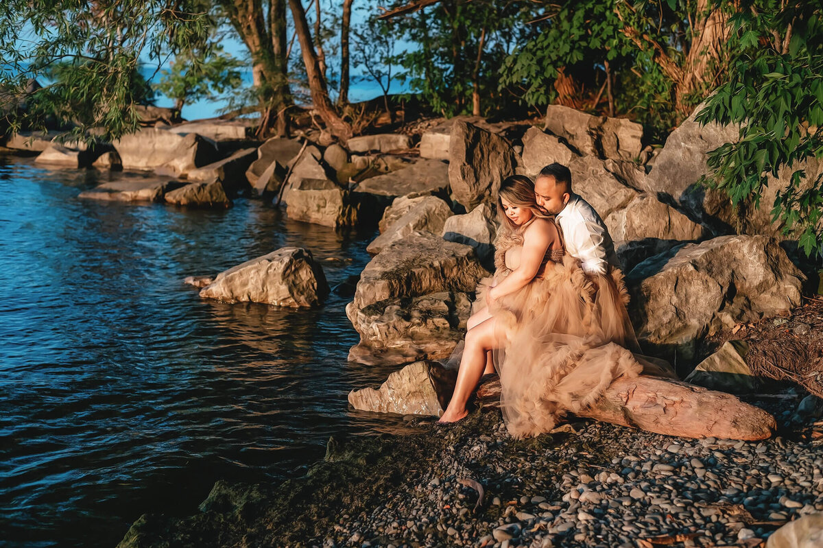 Couples cuddling on a rock at their pregnancy session at the lake.
