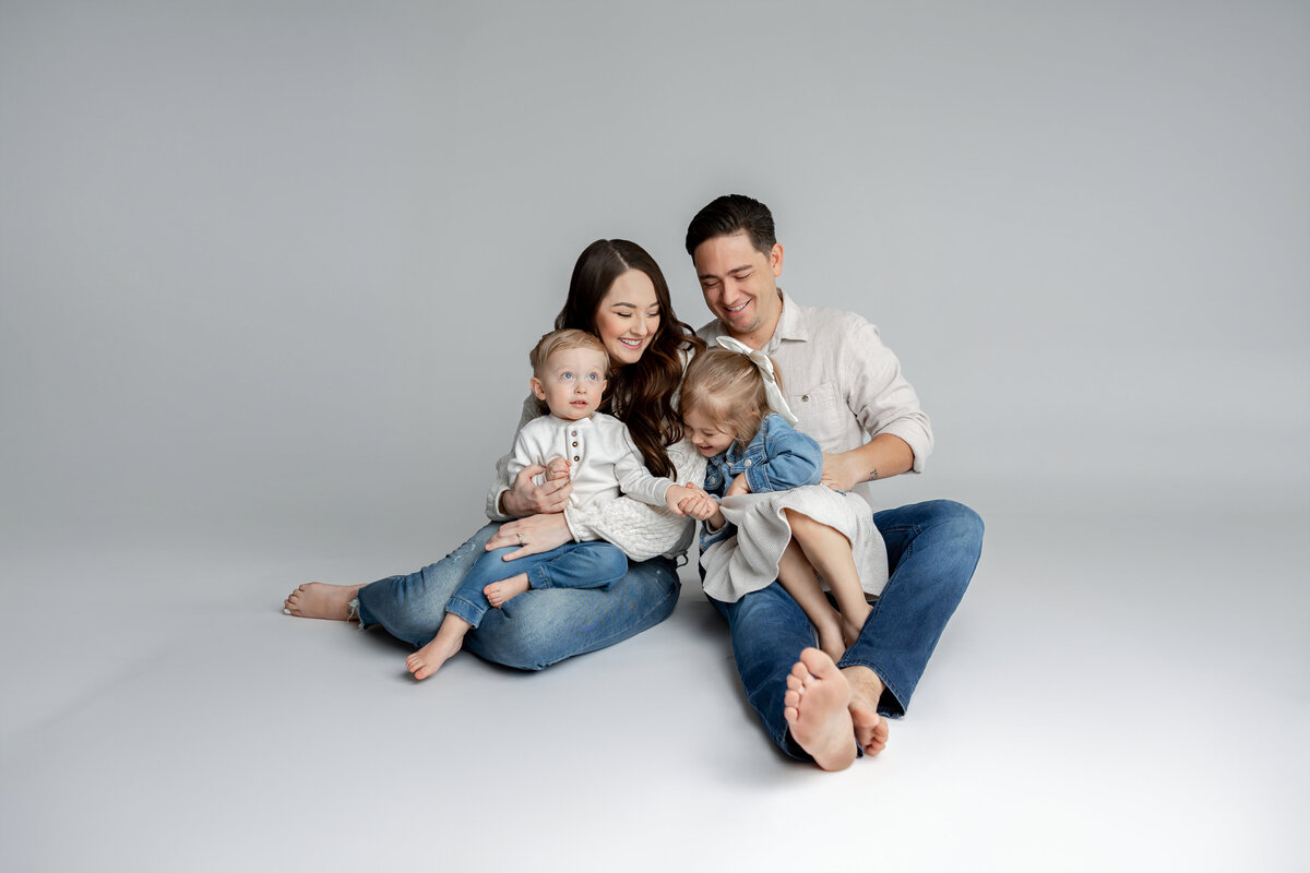 Family of four sitting on floor in studio, sharing a warm moment in denim and floral outfits. Photographed by Yucaipa family photographer Kaitlyn Dawn Photography.