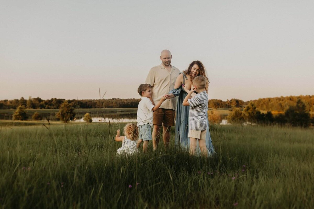 A family of five stands together in a grassy field near a lake at sunset, with two adults and three young children interacting during a family session in Warner Robins GA by Fire Family Photography.