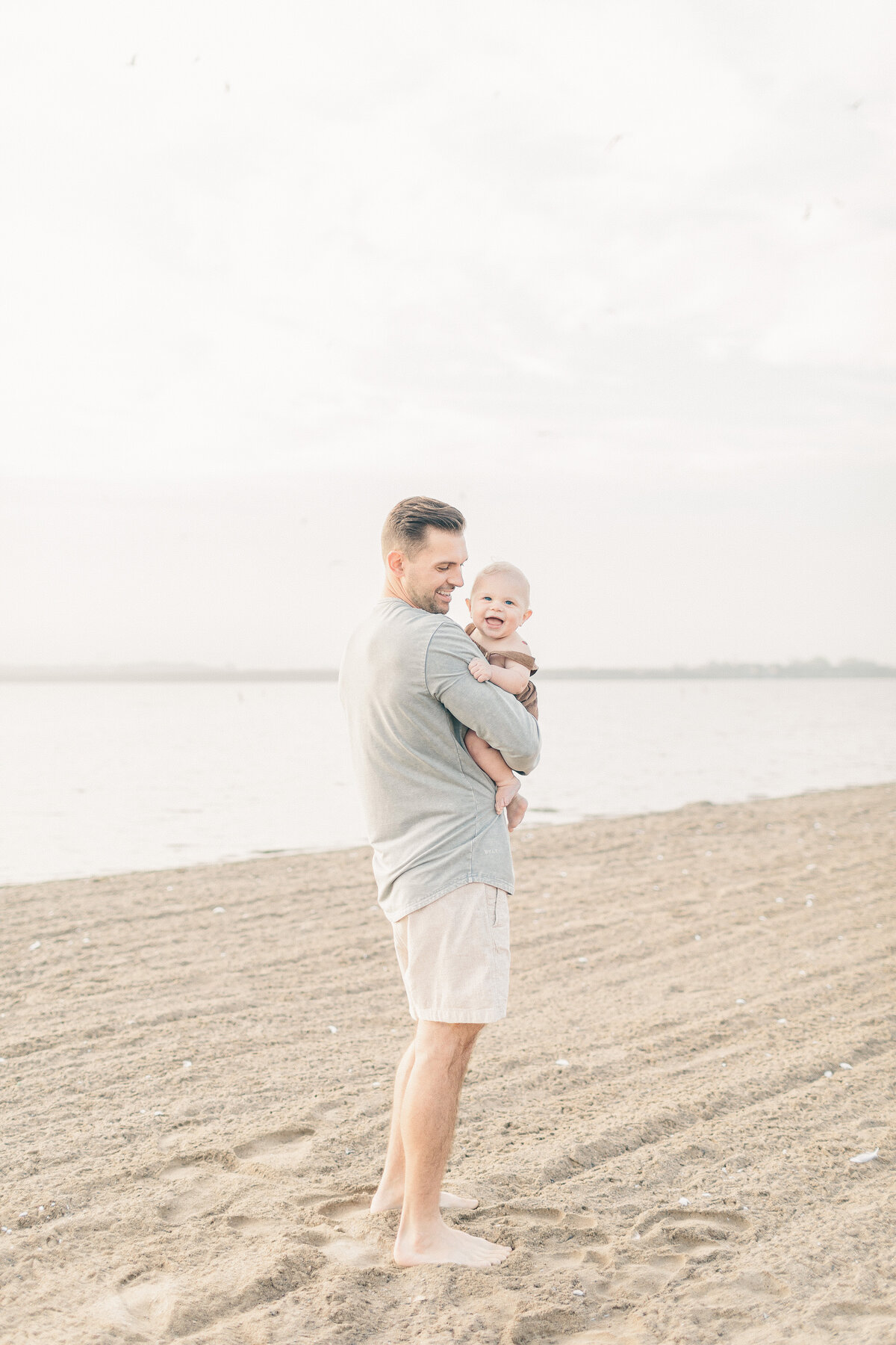 Dad holding his baby boy on the beach during a sunny 6-month milestone session at a Kansas City lake.