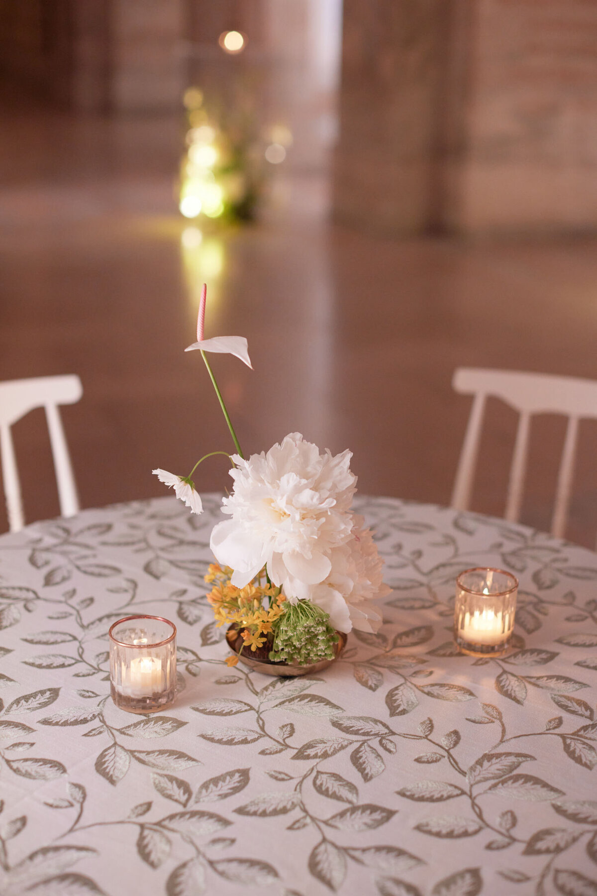 The photo was taken at the New York Public Library in New York City by photographer Shawn Connell. It features a round table with a floral-patterned tablecloth, showcasing a flower arrangement with white and yellow blooms at its center. Additionally, two lit votive candles in small glass holders flank the arrangement, creating a warm and inviting ambiance. In the background, blurred lights add a touch of elegance to the overall composition. Videographers Aaron Navak Films captured the moment beautifully.