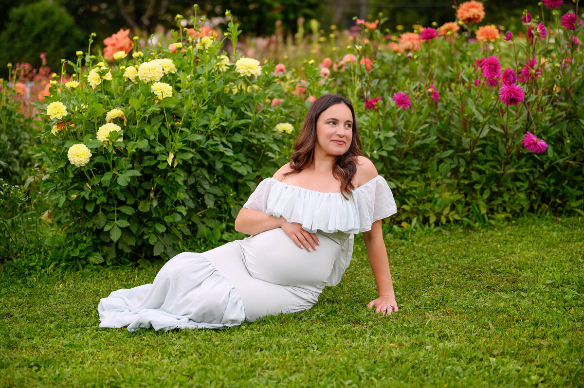 pregnant lady wearing a mint maternity dress sitting on the ground in front of flower for her outdoor maternity photography session in Burlington, Ontario