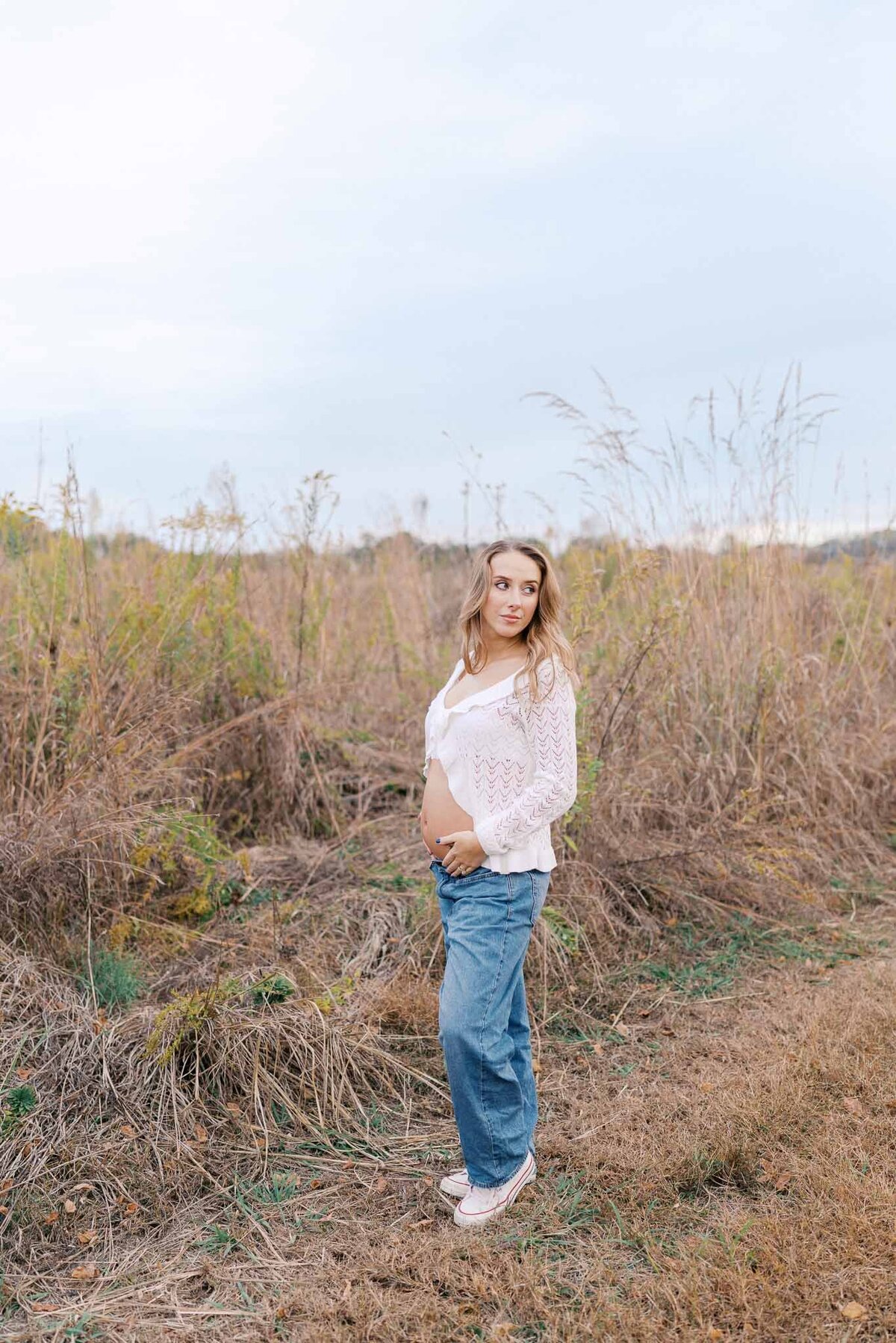 pregnant woman in white lace shirt holds belly in field during golden hour in knoxville tennessee
