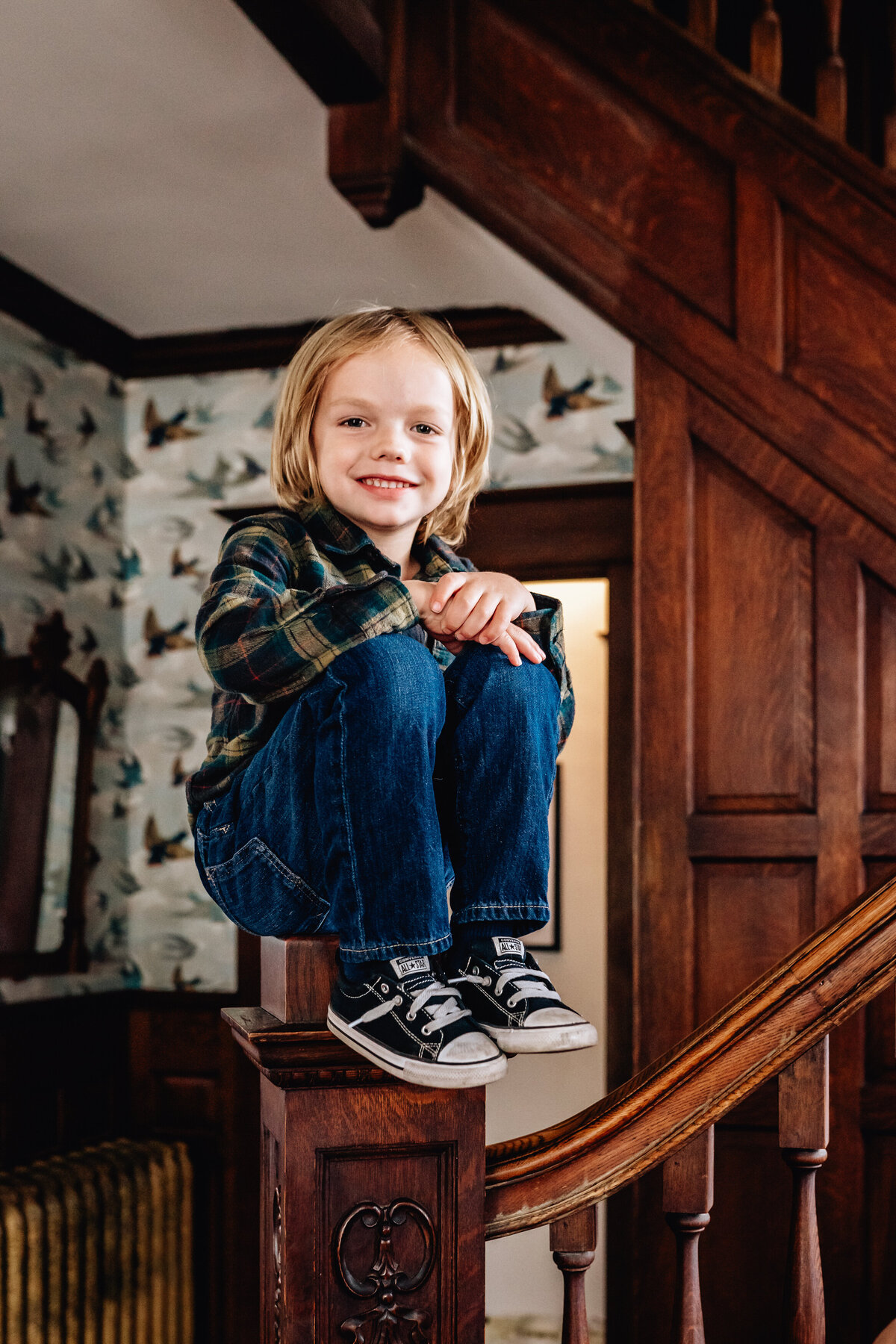 A young child sitting confidently on the bannister of a wooden staircase, smiling at the camera. The background shows wallpaper with birds and vintage decor.