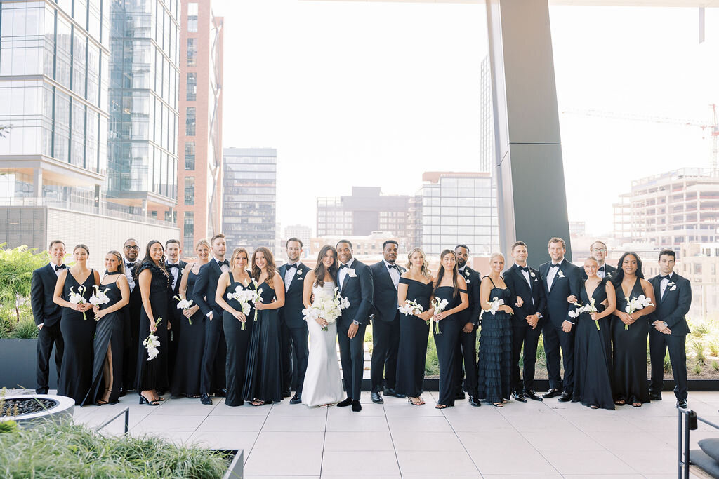 The bride, groom, and their wedding party dressed in black tie attire, posing on a rooftop with a city of Chicago skyline backdrop.