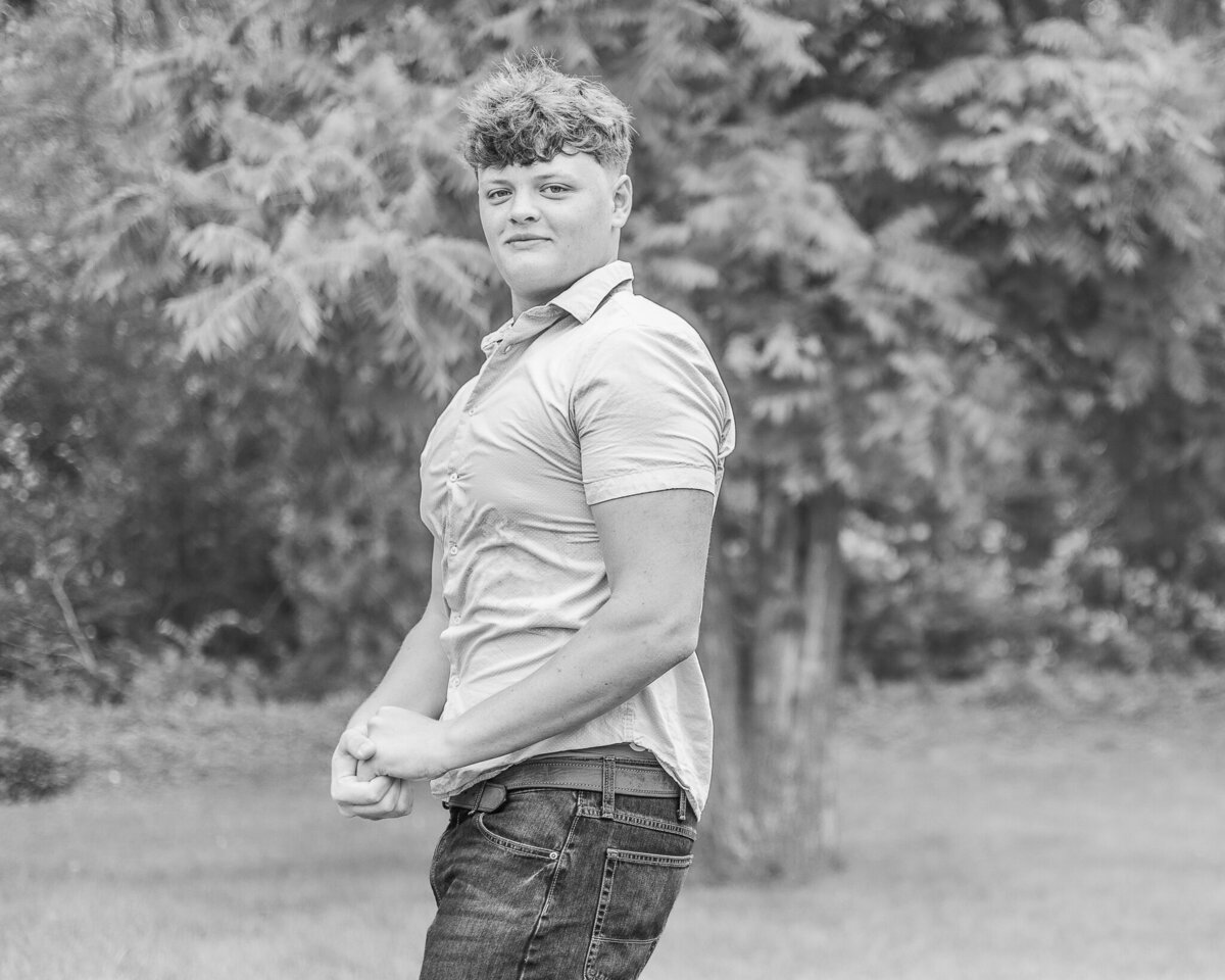 Black and white photo of oldest son standing in the grass with a tree line behind him turned to the side, flexing his muscles, looking at the camera with a soft smile, wearing jeans and short sleeve button up shirt.