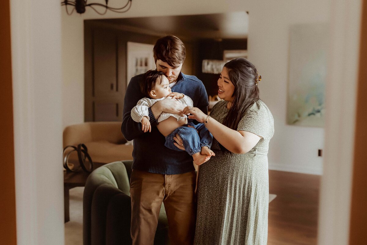 A man and woman stand close together in a living room, smiling while holding a baby. The room has neutral tones with a green armchair and wall art in the background.