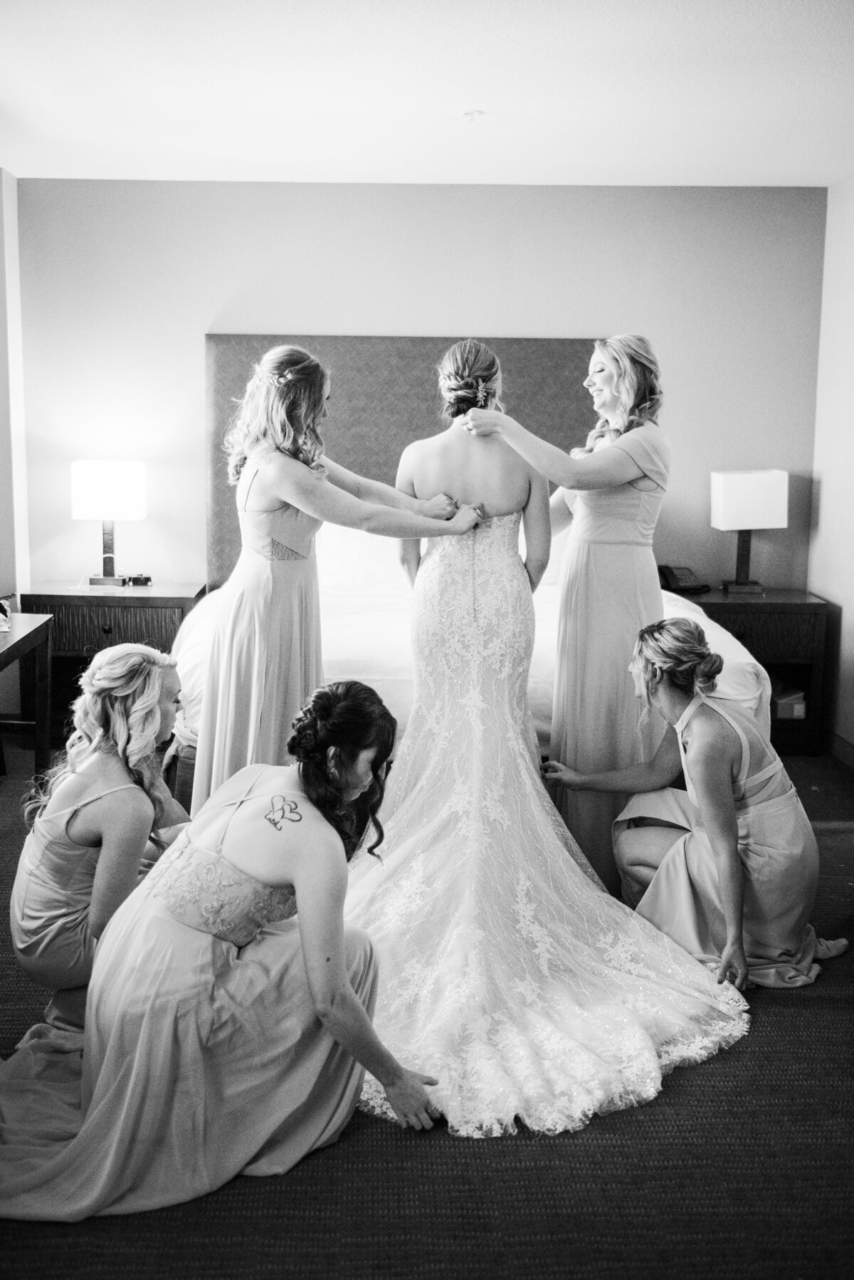 A bride faces away from the camera as all her bridesmaids adjust her wedding dress and train.