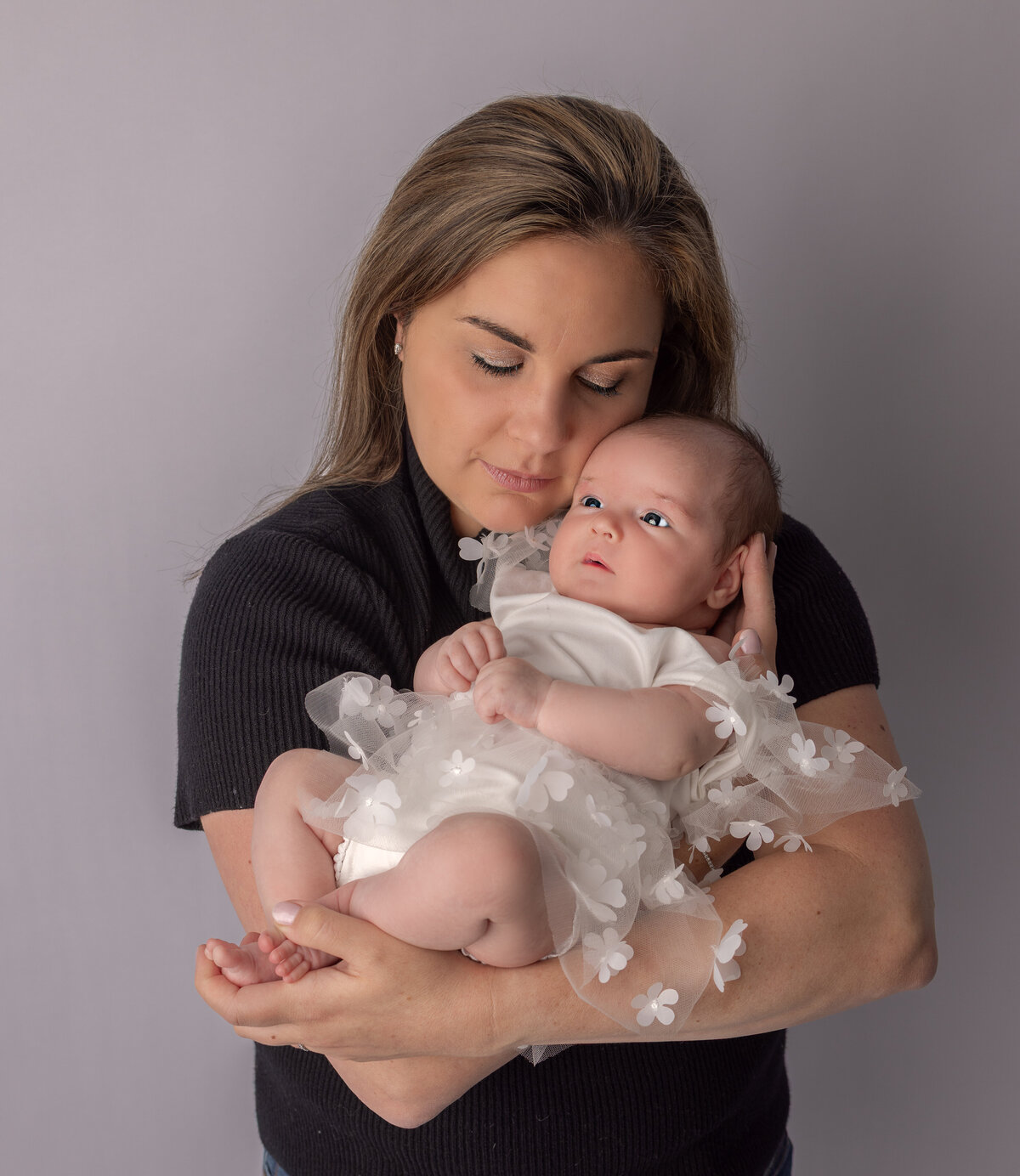 Mother lovingly holding her newborn baby girl dressed in a white floral outfit during a studio newborn session in NJ
