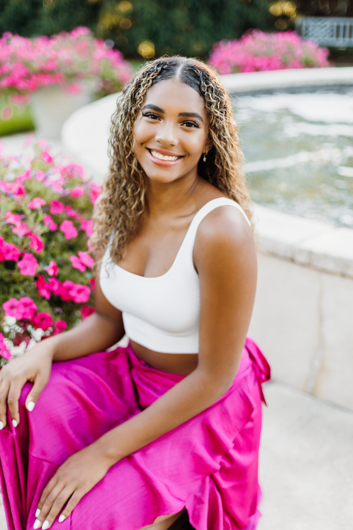 Senior girl in a white top and pink skirt posing in front of pink flowers and a fountain