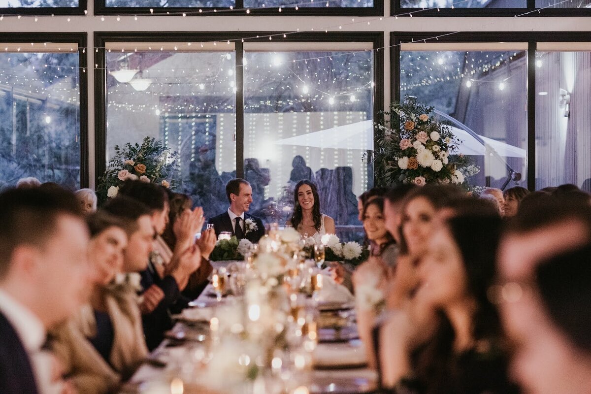 Bride and groom framed by floral pillar installation and guests at long table at their indoor wedding reception.