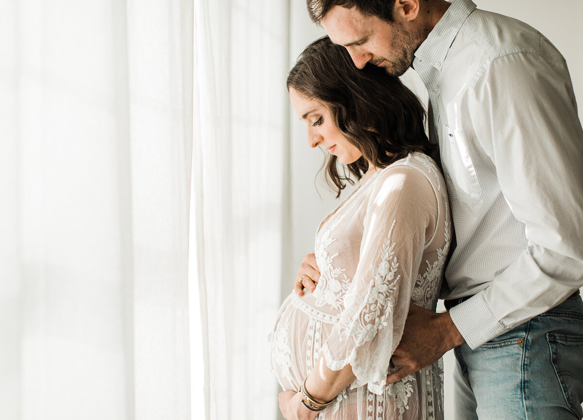 dad hugs mom from behind and rests his hands on her growing baby bump