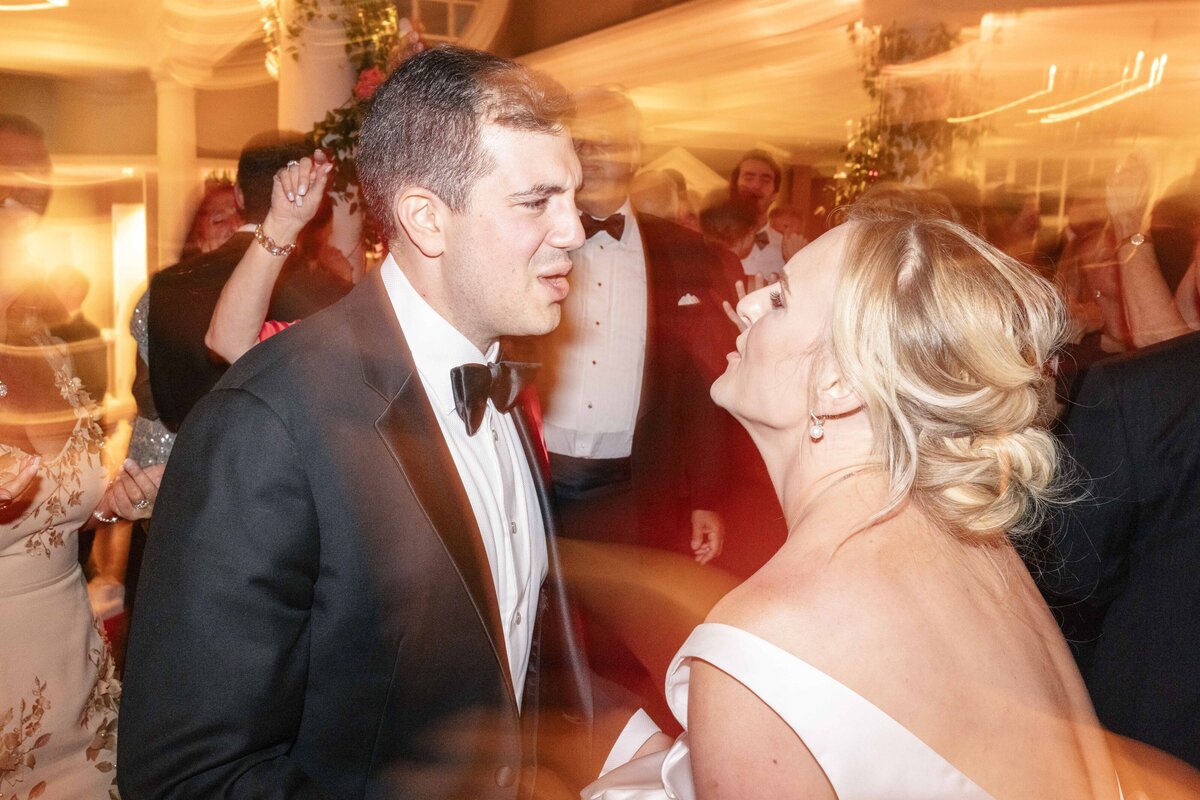 A man and woman in formal attire enjoy a lively event. The woman wears an off-shoulder white dress, and the man is in a black tuxedo. The background is blurred with warm lights, indicating movement and energy from the party.