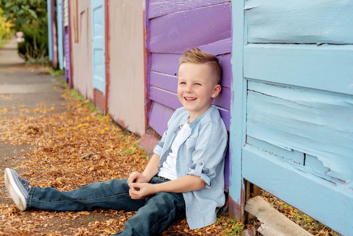 Little boy sitting on some fall leaves in front of a blue and purple fence made of old doors.