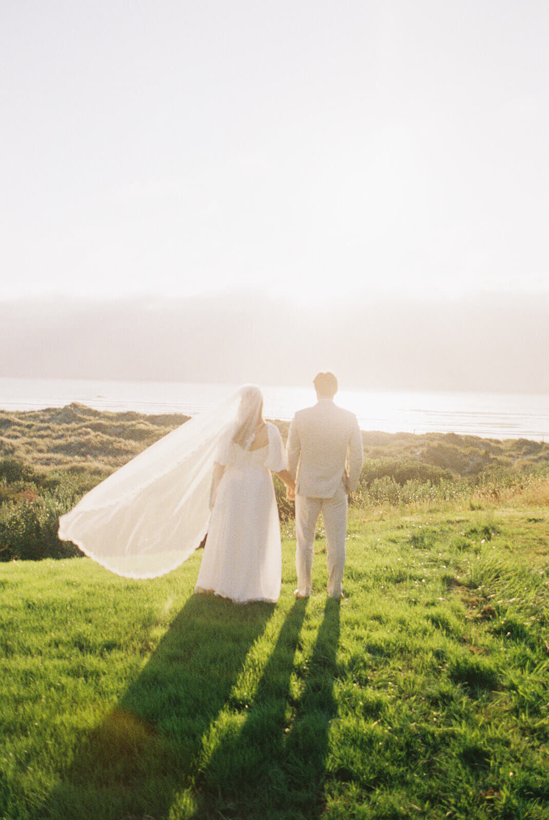 A bride and groom holding hands and facing away from the camera with the ocean in the background.