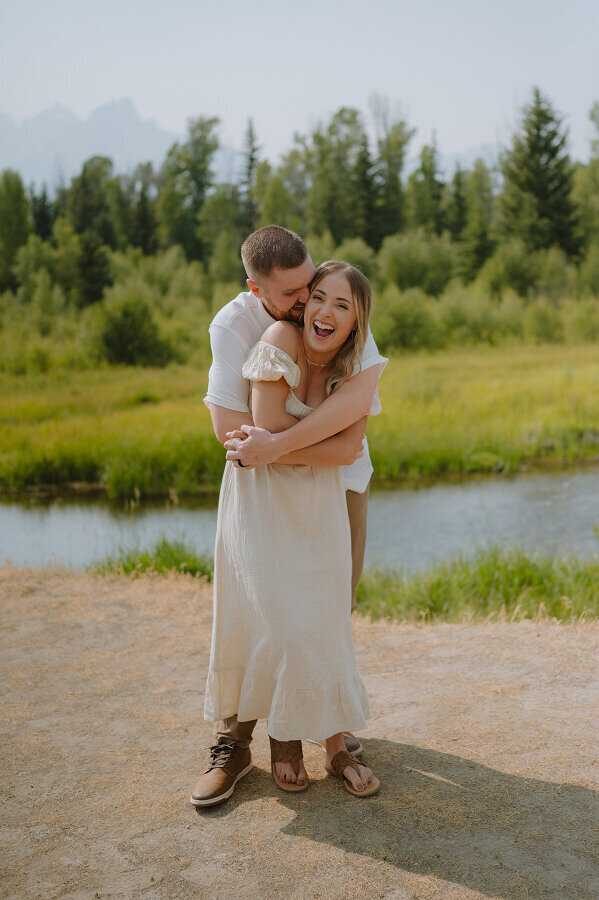 A man holds his wife from behind in a bear huge as she smiles big. Behind them is a river and lots of trees.