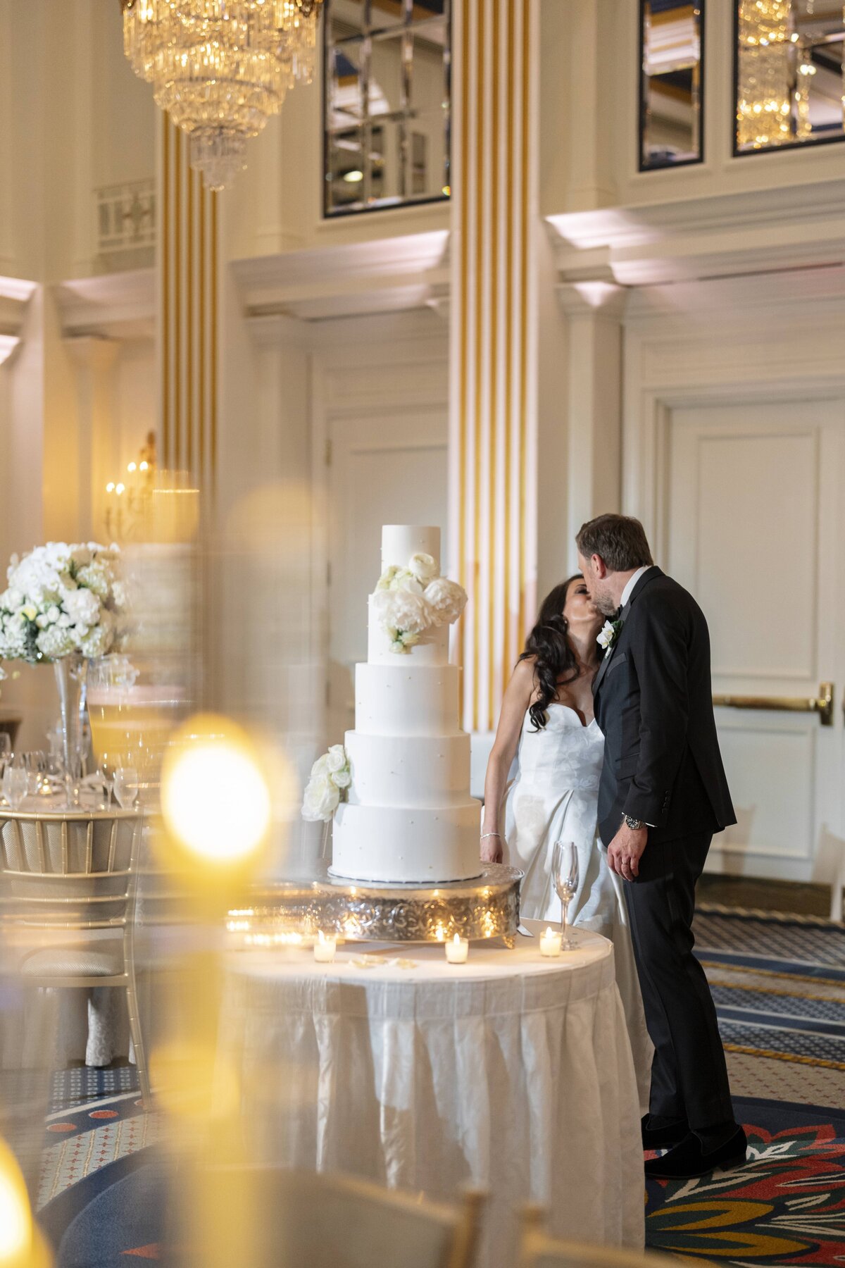 A couple in wedding attire kisses beside a tall, white tiered wedding cake in an elegant venue. The room is decorated with white flowers, candles, and gold accents, creating a romantic atmosphere.