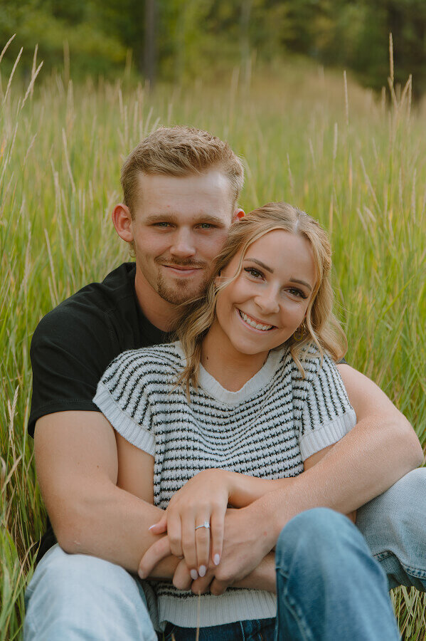 Close up portrait of a couple sitting in a tall grassy field. The man is holding his fiancé between his legs.